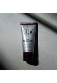 Grå tub med röda accenter, märkt "2-i-1 Hudfuktare & Skäggbalsam" av American Crew. Fliplock på basen, 100 mL.