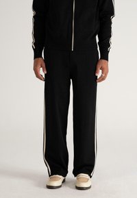 Male wearing black track pants with cream side stripes, matching black zipped jacket, and cream woven loafers, standing on light background.
