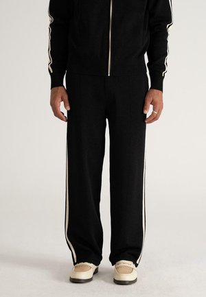 Male wearing black track pants with cream side stripes, matching black zipped jacket, and cream woven loafers, standing on light background.