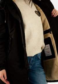 Chaqueta exterior negra aislante con forro de piel sintética; suéter de punto beige por debajo con un parche en forma de corazón de leopardo; jeans azules.