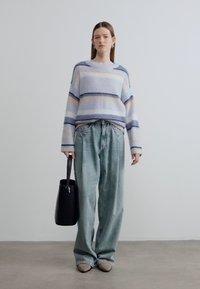 Pull légèrement rayé bleu et crème, associé à un jean ample et clair. Tenant un sac fourre-tout noir à finition lisse.