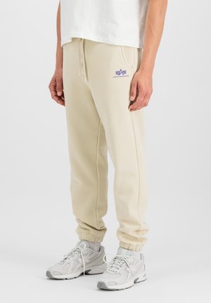 Person, die beige Alpha Industries Sweatpants und weiße Sportschuhe trägt, steht vor einem schlichten hellen Hintergrund.