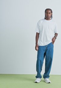 Hvid t-shirt med korte ærmer, blå baggy jeans og hvide sneakers. Baggrunden er lys grå med et grønt gulv. En enkel og afslappet påklædning.