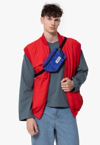 Gilet imbottito rosso senza maniche sopra una maglietta grigia a maniche lunghe, abbinato a una borsa a spalla in corduroy blu. I jeans di denim completano l'outfit.