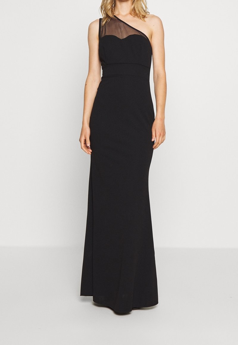 Femme portant une longue robe de soirée noire une épaule, avec un panneau transparent sur l'épaule, debout devant un fond uni.