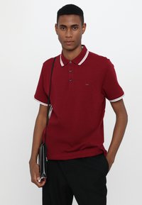 Burgunderfarget polo-skjorte med hvite striper på krage og mansjetter, tre-knappers knapping, og liten logo. Kombinert med svarte bukser og clutch.