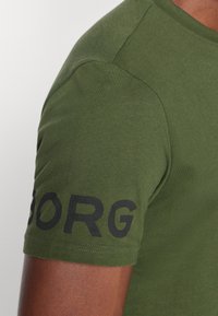 Olivenfarget bomull t-skjorte med svart tekst på venstre erme. Glatt tekstur med klassisk rund hals og korte ermer.
