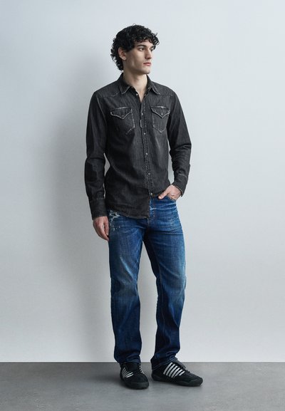 Νεαρός με σγουρά σκούρα μαλλιά, φοράει μαύρο πουκάμισο με κουμπιά, μπλε τζιν και μαύρα αθλητικά παπούτσια, στέκεται μπροστά σε έναν απλό φωτεινό τοίχο.