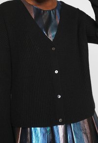 Černý žebrovaný pletený cardigan s výstřihem do V, zapínáním na knoflíky a dlouhými rukávy, doplněný o lesklou, vzorovanou metalickou sukni.