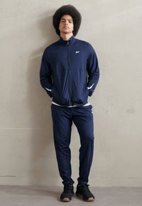 Chándal de color navy hecho de tela ligera, que incluye una chaqueta con cremallera y pantalones ajustados. Presenta detalles en blanco y zapatos negros.
