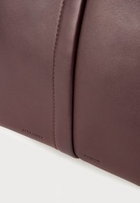 Brun lædertaske med en glat overflade, kontraststing, og prægede branding på siden, med et struktureret, minimalistisk design.