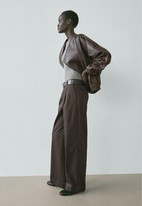 Große Person mit kurzen Haaren, die eine kurze braune Lederjacke, ein graues eng anliegendes Oberteil, weite braune Hosen und schwarze Schuhe trägt, seitlich stehend.