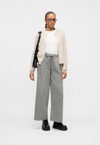 Νέα γυναίκα με πλεξούδες, που φορά γυαλιά, μπεζ ζακέτα, λευκό πουκάμισο, γκρι φαρδιά παντελόνια, μαύρα μποτάκια και μαύρη τσάντα ώμου.