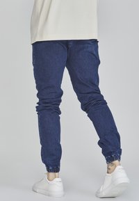 Denimjoggers med en avsmalnande passform, elastiska muddar och en mörkblå tvätt, tillsammans med vita sneakers mot en ljus bakgrund.