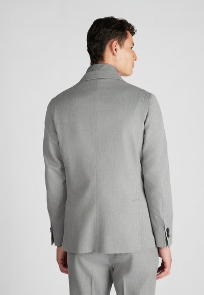 Uomo vestito con un completo grigio chiaro e pantaloni abbinati, visto da dietro su uno sfondo chiaro e semplice.