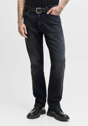 Hombre con vaqueros negros, cinturón negro, zapatos de cordones negros, con brazos tatuados visibles, mano en el bolsillo, de pie contra un fondo liso.