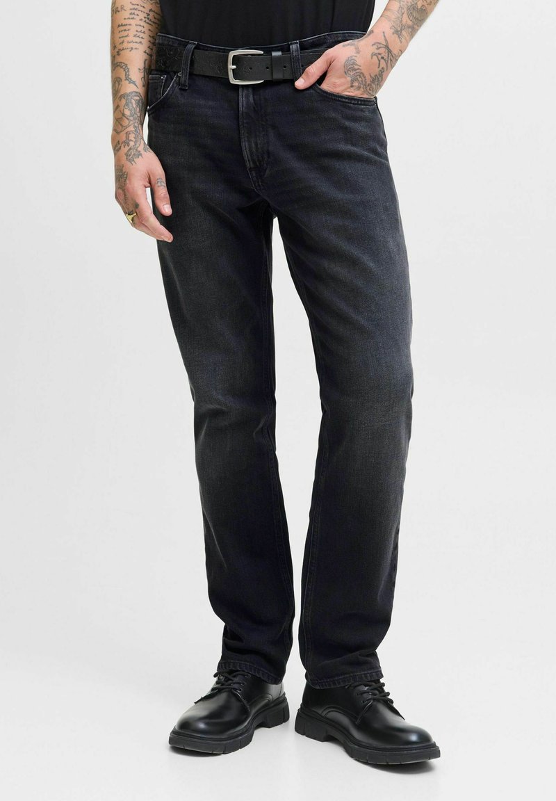 Hombre con vaqueros negros, cinturón negro, zapatos de cordones negros, con brazos tatuados visibles, mano en el bolsillo, de pie contra un fondo liso.