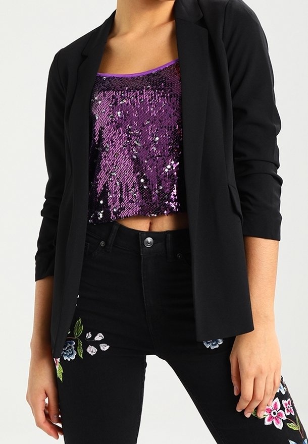 Blazer noir sur un crop top violet à sequins, associé à un jean noir orné de broderies florales colorées à l'avant.