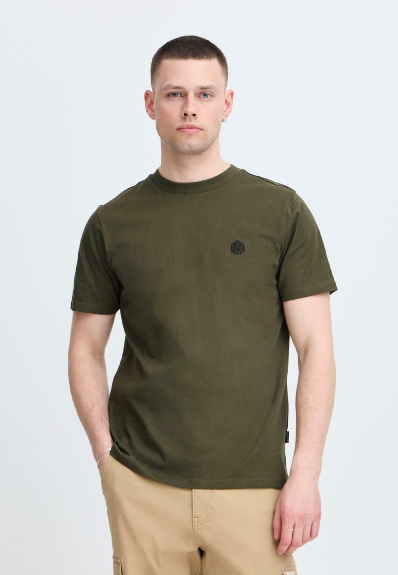 Tricou din bumbac de culoare verde măslinie, cu mâneci scurte și guler rotund, având un logo hexagonal negru pe piept. Textură netedă și croială lejeră.