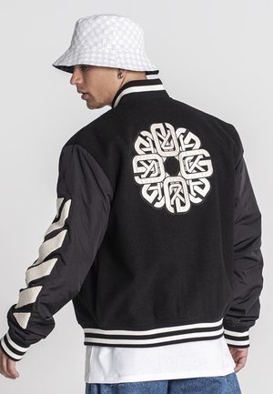 Black bomber jacket with embroidered white logo on the back, striped cuffs, and contrasting black sleeves. Paired with a patterned bucket hat.