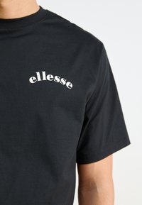 Camiseta de algodón negra con mangas cortas que presenta un logo curvado en blanco de "ellesse" en el lado izquierdo del pecho. Textura suave, corte clásico.