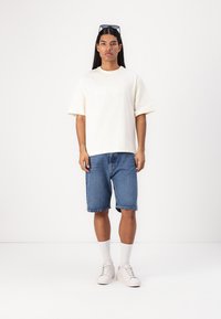 Kremfarget kortermet t-skjorte, blå denimshorts, hvite sneakers og hvite knesokker. Modellen står mot en ensfarget hvit bakgrunn.