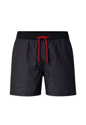 Skjorteløs mand iført sorte mønstrede badeshorts med rød snor, sorte klipklappere og futuristiske ombøjede solbriller, stående afslappet.
