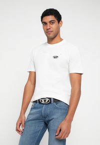 Bela majica s kratkimi rokavi z majhnim grafičnim motivom očesa na prsih. Nošena z modrimi jeans hlačami in črnim pasom s srebrno sponko.