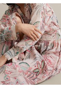 Personne tenant son bras, portant une robe rose pâle à motifs floraux avec des manches longues et un motif botanique détaillé.