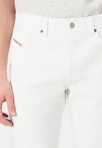 White denim jeans feature a classic five-pocket design, silver hardware, and a small label on the front pocket with red accents.