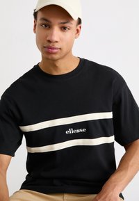 Juodas medvilnės marškinėliai su dviem horizontaliomis baltomis juostelėmis ir "ellesse" logotipu. Trumpomis rankovėmis ir apvaliu iškirpimu. Lygaus paviršiaus.