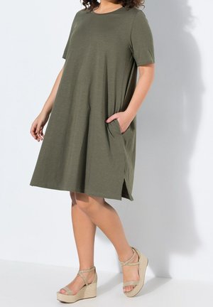 Plus size kvinde iført olivengrøn knælængde kjole med korte ærmer og sidelommer samt beige kilehøjhælte sandaler, stående mod hvid baggrund.