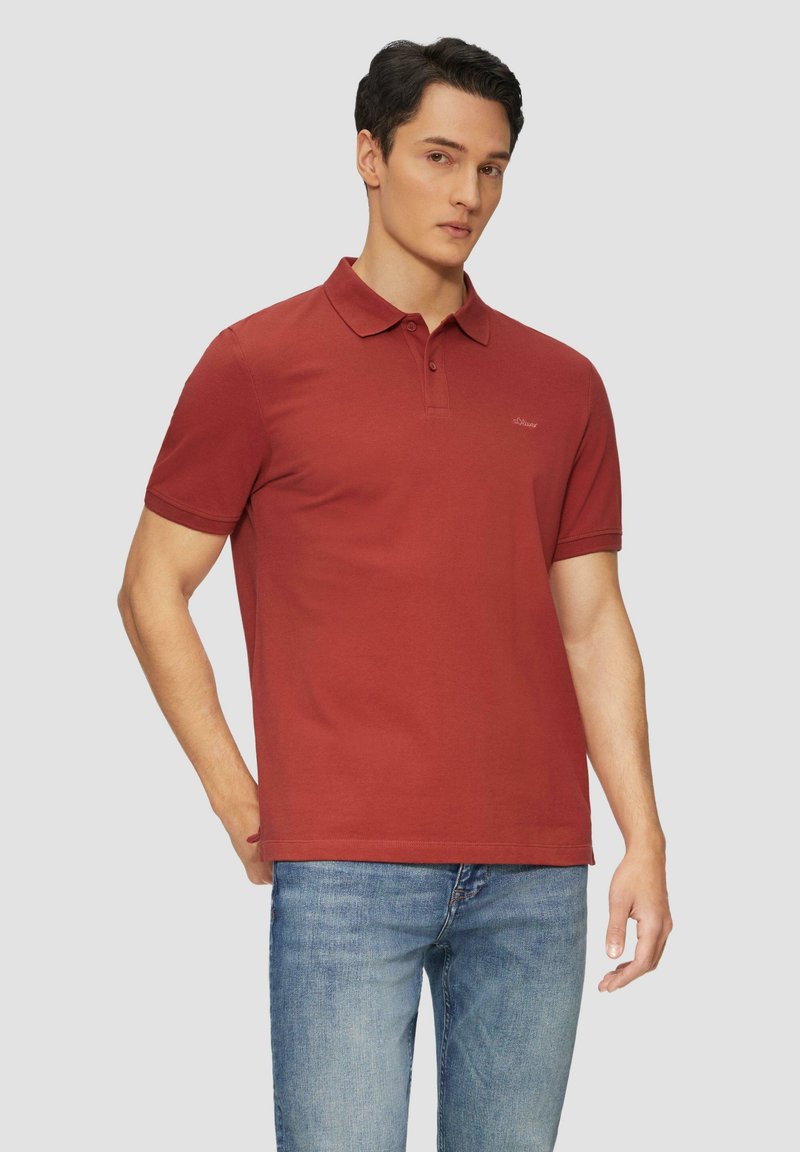T-shirt polo rouge en coton, avec un col et des manches courtes. Logo distinctif sur la poitrine. Porté avec un jean bleu.