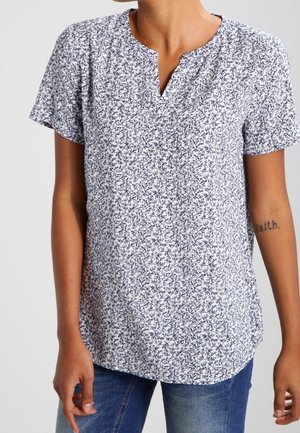 Persona que lleva una blusa blanca de manga corta con un estampado floral negro y jeans azules, mostrando un tatuaje que dice "faith" en el antebrazo izquierdo.