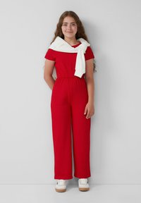 Combinaison rouge côtelée à manches courtes, taille ceinturée, jambes larges, accompagnée d'un pull blanc drapé sur les épaules et de baskets blanches.