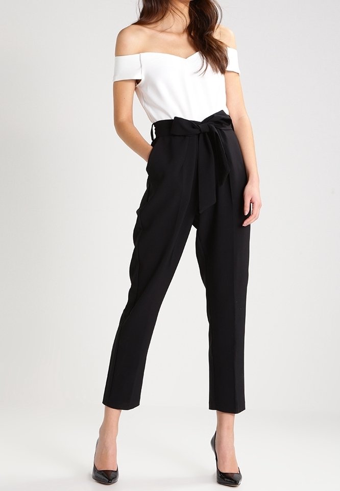 Femme portant un top blanc épaules dénudées, un pantalon paperbag noir taille haute avec une ceinture nouée, et des talons pointus noirs, debout devant un fond uni.