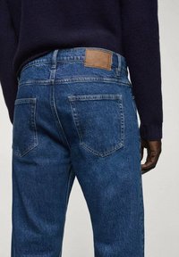 Jeans de mezclilla azul con una textura suave, que presentan un parche de cuero marrón en la cintura y dos bolsillos traseros con costuras curvadas.