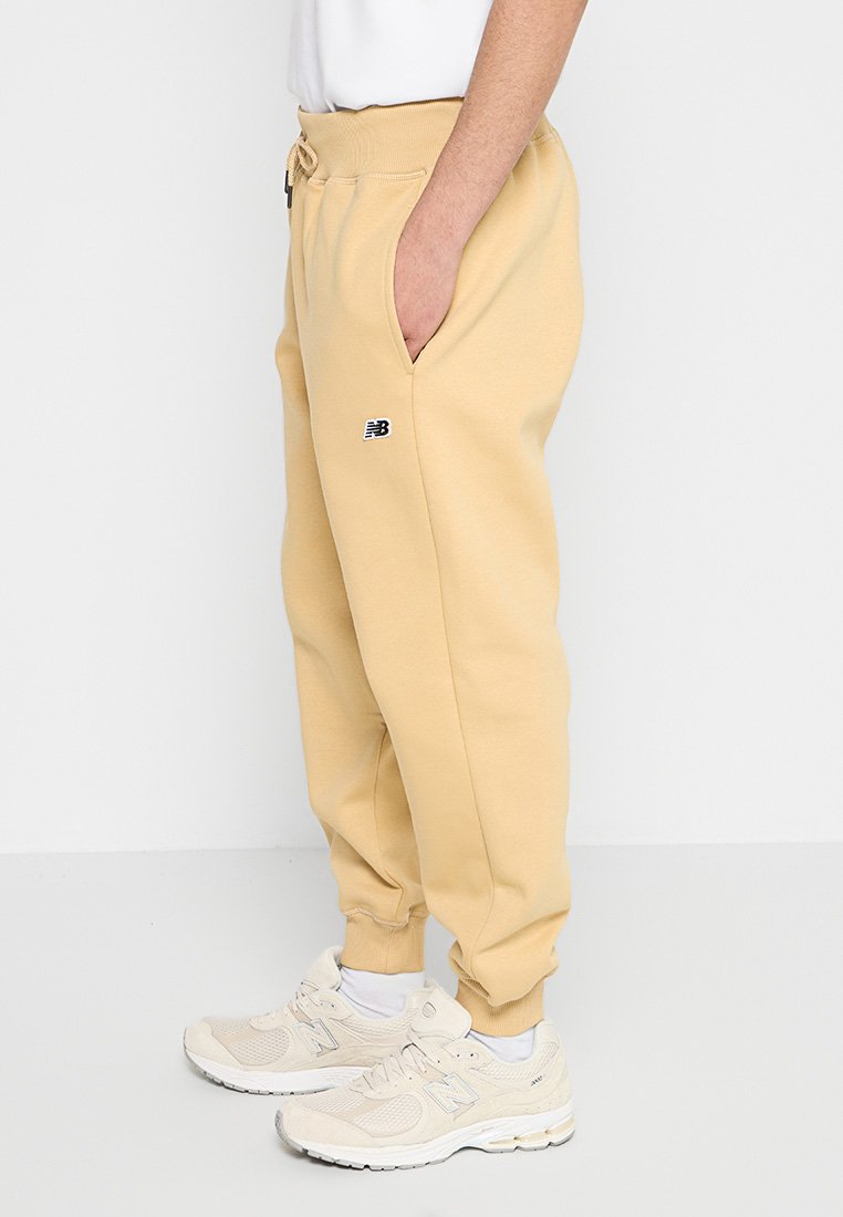 Pantalones deportivos beige con cintura elástica y cordón ajustable, que cuentan con bolsillos laterales y un pequeño logotipo negro en el muslo izquierdo.
