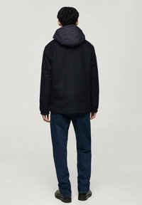 Černá bunda s šedou kapucí, strukturovaná látka a přední kapsy, v kombinaci s modrými džíny a černými botami, pohled zezadu.