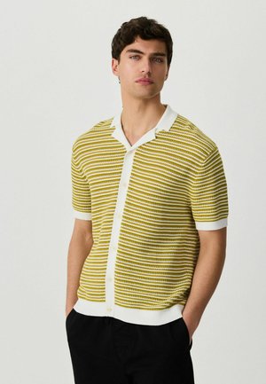 Ung mand med mørkt krøllet hår iført en gul og hvid stribet kortærmet skjorte med knapper, med hænderne i lommerne på sorte bukser.