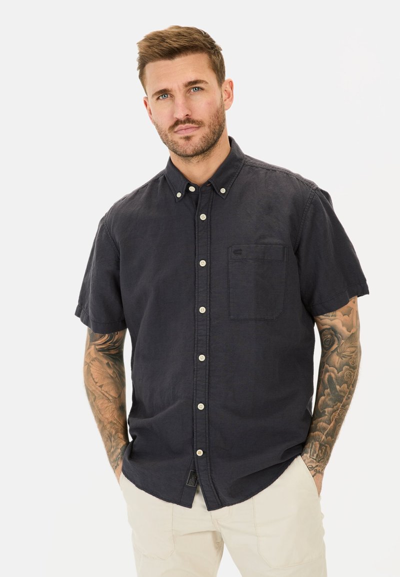Korte mouwen, marineblauw knoopshirt met een borstzak, gemaakt van geweven stof; heeft witte knopen en een ronde onderkant.