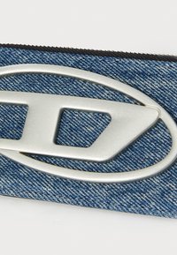 Læren lommebok i denim med et stort sølvfarget metalllogo, teksturert materiale og en glidelås langs toppen for enkel tilgang.