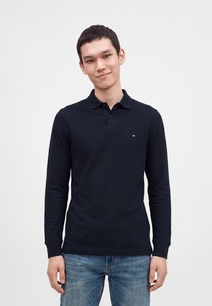 Jeune homme aux cheveux courts et foncés, portant un polo à manches longues foncé et un jean bleu, debout devant un fond clair et uni.