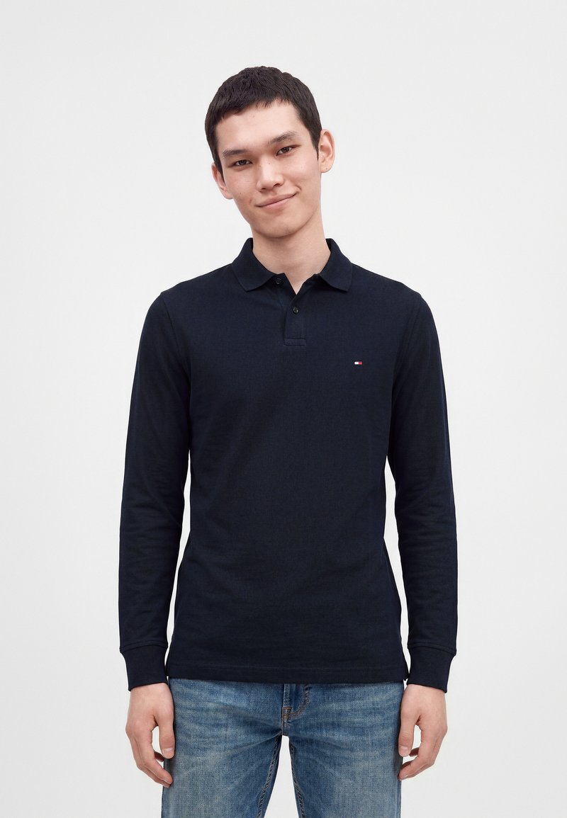 Jeune homme aux cheveux courts et foncés, portant un polo à manches longues foncé et un jean bleu, debout devant un fond clair et uni.