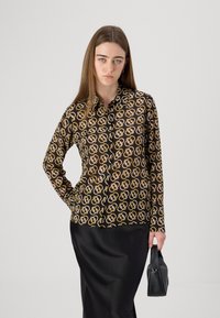 Maglietta nera a maniche lunghe con un motivo interlocking giallo e bianco ripetuto. Il modello tiene in mano una piccola borsa nera, abbinata a una gonna nera.