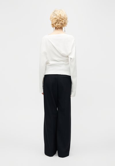 Άτομο με κοντά, σγουρά ξανθά μαλλιά που φοράει λευκό μακρυμάνικο τοπ και μαύρο παντελόνι με φαρδύ πόδι, στέκεται γυρισμένο προς τα πίσω σε απλότερο φόντο.