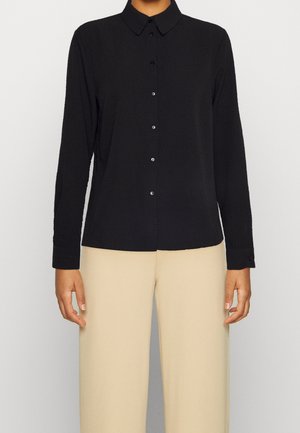 Schwarzes Langarmshirt mit Button-Down-Kragen, dezent strukturiertem Stoff und passenden Knöpfen, kombiniert mit beigen Hosen.