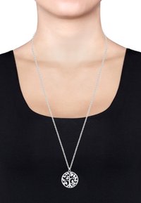 Sølvhalskæde med en rund vedhæng, der har et indviklet trædesign. Kæden er delikat og hviler elegant mod en mørk bluse.