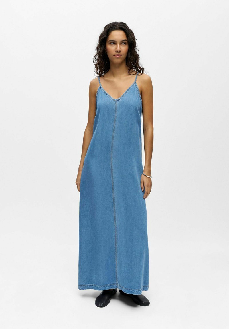 Femme aux cheveux bouclés portant une robe longue en jean bleu sans manches et des chaussures noires, debout devant un fond blanc uni.