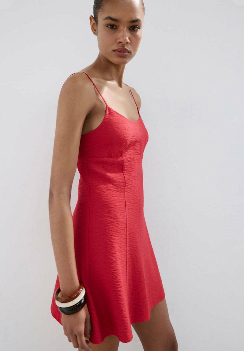 Femme portant une robe rouge texturée sans manches à fines bretelles, avec trois bracelets larges à son poignet gauche, sur un fond uni.